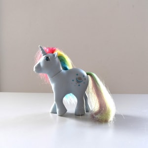 Op de afbeelding: Een lichtblauw eenhoornspeelgoed met een regenboogmanen en -staart. De eenhoorn heeft een witte hoorn en blauwe ogen. Het speelgoed heeft een ontwerp op de achterkant en staat op een wit oppervlak.