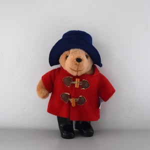 Op de afbeelding: Een Paddington Bear knuffel, gekleed in een marineblauwe hoed, een rode jas met houten knevelsluitingen en zwarte laarzen. De beer heeft een vriendelijke uitdrukking en is een klassiek kinderspeelgoed.