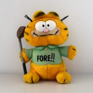 May include: A plush Garfield toy dressed for golf. The orange cat has black stripes, large white eyes, and a pink nose. He wears a green shirt with "FORE!!" printed on it and holds a golf club. The toy is on a white surface.