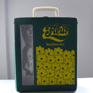 May include: A dark green box with a white handle and a clear window. The box has yellow floral patterns and the text "Fleur" and "You'll love her" in yellow. The box is sitting on a white surface.