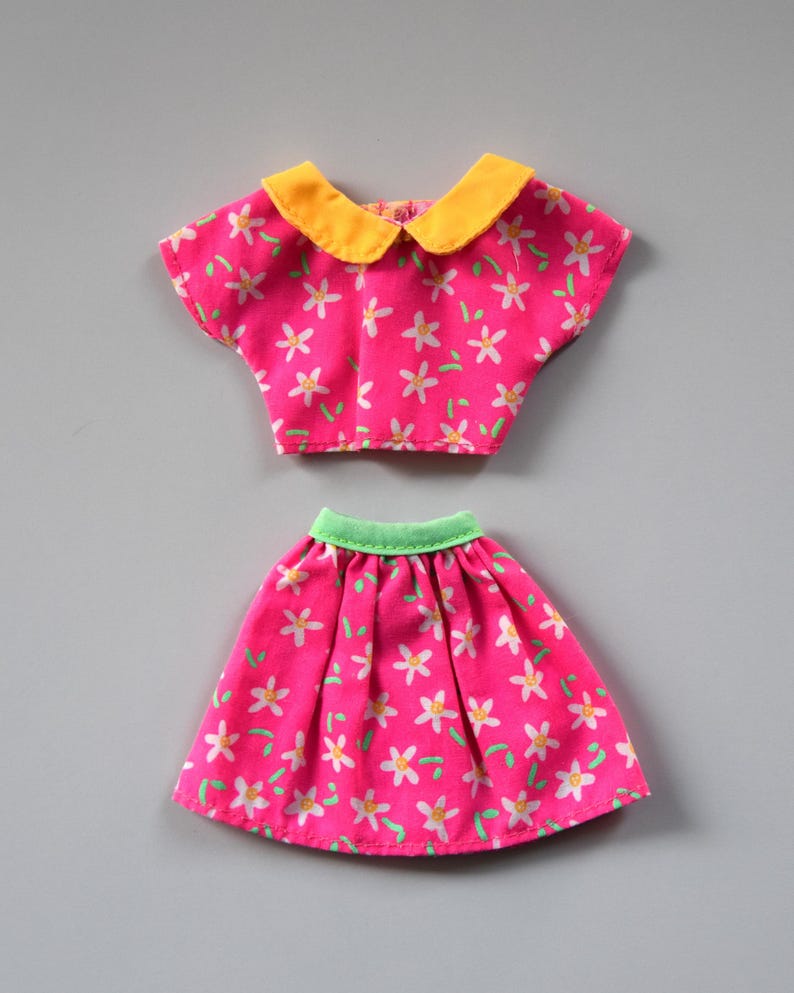 May include: A pink and white floral print doll outfit with a yellow collar and green waistband.