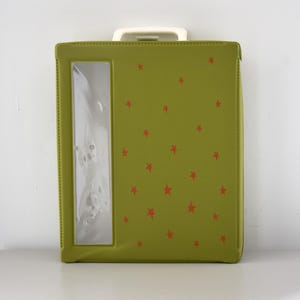 May include: A green binder with a clear window and a white handle. The binder is decorated with small red stars. The binder is closed and appears to be made of a durable material.
