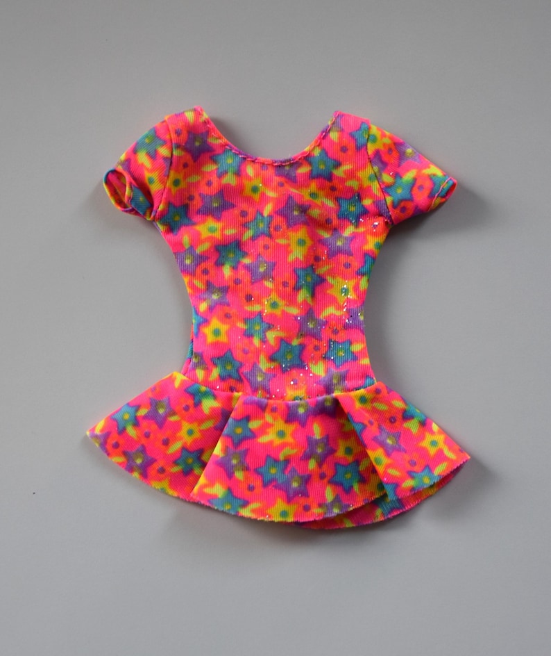 May include: A pink leotard with a multi-colored star print. The leotard has short sleeves and a ruffled skirt.