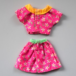 May include: A pink and white floral print doll outfit with a yellow collar and green trim. The outfit includes a short-sleeved top and a skirt.