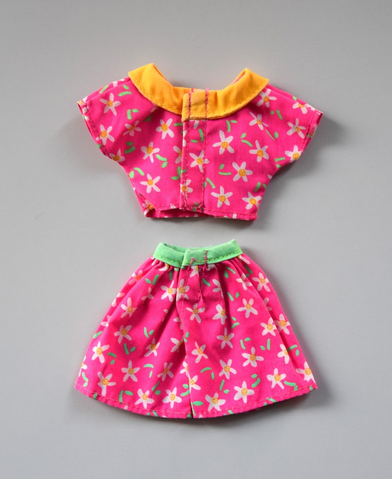 May include: A pink and white floral print doll outfit with a yellow collar and green trim. The outfit includes a short-sleeved top and a skirt.