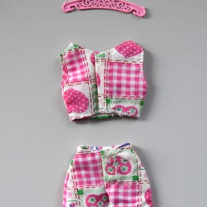 May include: A pink and white gingham doll dress with a ruffled hem and a matching pink top. The top has a heart and flower pattern.