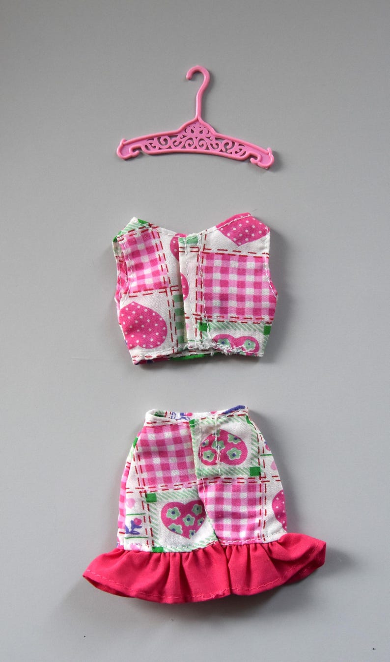 May include: A pink and white gingham doll dress with a ruffled hem and a matching pink top. The top has a heart and flower pattern.