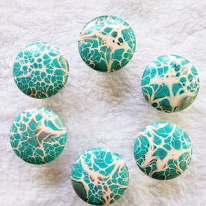 May include: Six teal and white round knobs with a marbled pattern. The knobs are arranged in a circle on a white fuzzy surface.