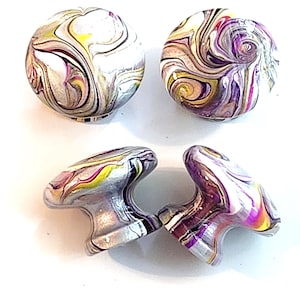 May include: Four cabinet knobs featuring a marbled pattern with swirls of purple, yellow, white, and black. Two knobs are shown from a top-down perspective, while the other two are positioned on their sides to display the flared base and stem.