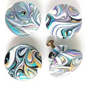 May include: Four round cabinet knobs featuring a swirling marbled pattern in shades of white, teal, purple, black, and gold. Each knob has a smooth, glossy finish. One knob is shown from the side, revealing a metal screw attached to the back.