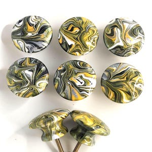 May include: Eight round cabinet knobs featuring a marbled pattern in shades of yellow, black, white, and metallic gold. The knobs have a glossy finish and a smooth, rounded surface. Two of the knobs are shown from the side, revealing a flared base and a threaded metal screw for installation.