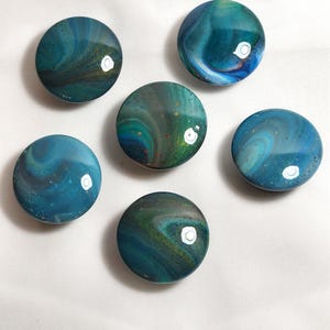 May include: Five circular cabinet knobs featuring a glossy, poured paint finish in shades of teal, deep blue, and forest green with subtle gold glitter accents. Each knob displays a unique, swirling marbled pattern with a smooth, reflective surface.