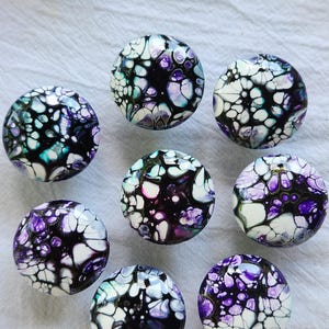 May include: A collection of seven round cabinet knobs featuring a fluid acrylic pour design. Each knob displays a marbled pattern of black, white, purple, and teal colors, creating a glossy, cell-like effect across the smooth, domed surfaces.
