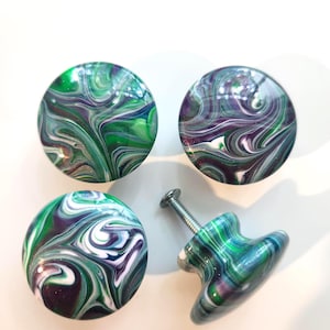 May include: Four round cabinet knobs featuring a swirling marbled pattern in shades of emerald green, deep purple, and white. Each knob has a smooth, glossy finish and a metallic screw attached to the back of one knob for installation purposes.