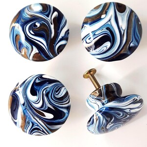 May include: Four round cabinet knobs featuring a swirling marbled pattern in shades of navy blue, light blue, white, and metallic gold. Each knob has a smooth, glossy finish and includes a threaded metal screw attached to the back for installation.