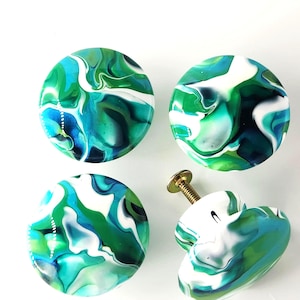 May include: Four round cabinet knobs featuring a marbled pattern of swirling green, teal, white, and dark blue colors. Each knob has a smooth, glossy finish and a metallic screw post attached to the back for installation on furniture doors.