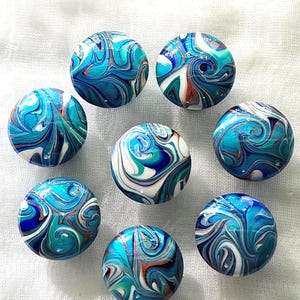 May include: Seven circular knobs featuring a marbled pattern of swirling blue, turquoise, white, and copper colors. Each knob has a smooth, glossy finish with a distinct, unique arrangement of colors that blend together in fluid, organic shapes.