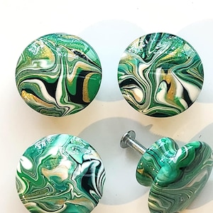 May include: Four round cabinet knobs featuring a marbled pattern in shades of emerald green, white, black, and metallic gold. Each knob has a smooth, glossy finish with a swirling design. One knob is shown from the side, revealing a metal screw.