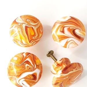 May include: Four round cabinet knobs featuring a swirling marbled pattern in shades of orange, white, and gold. Each knob has a smooth, glossy finish with a metallic screw attached to the back of one piece for installation on furniture.