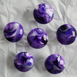 May include: Six round decorative knobs with a glossy finish. The knobs feature a swirling pattern of purple, white, and silver colors. The knobs are arranged on a white, textured surface.