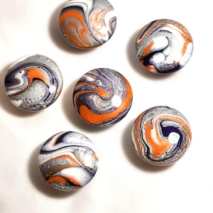 May include: A set of five circular buttons featuring a marbled pattern with swirls of orange, deep purple, white, and metallic silver. Each button has a smooth, glossy finish with a distinct, unique color distribution across the rounded surface.