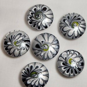 May include: Six round, silver-colored glass cabochons with black and white swirl patterns and a green center. The cabochons are arranged in a circle on a white background.