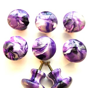 May include: A set of eight round cabinet knobs featuring a swirling marbled pattern in shades of purple, white, and black. Each knob has a smooth, glossy finish and includes a metal screw attached to the base for installation on furniture.