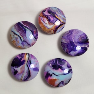 May include: Five round, purple, white, and gold abstract painted stones. The stones have a marbled, swirled effect.