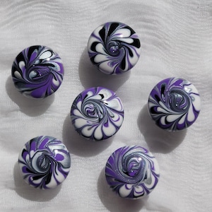 May include: Six round decorative cabinet knobs with a swirling design in shades of purple, black, white, and silver. The knobs are arranged on a white fabric surface, showcasing their unique, artistic patterns.
