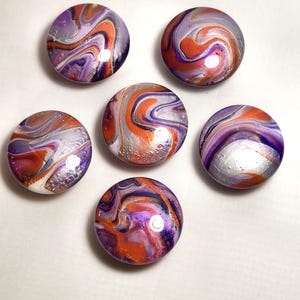 May include: A set of five circular cabinet knobs featuring a glossy, poured paint finish with swirling patterns of vibrant orange, deep purple, and metallic silver. Each knob has a smooth, rounded surface with reflective highlights and distinct color blends.