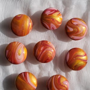 May include: Eight round, marble-like beads in shades of orange, pink, and gold. The beads have a swirled pattern, with gold glitter accents. The beads are arranged on a white, textured surface.