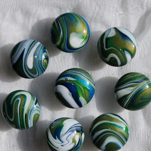 May include: Nine glass marbles with swirling patterns of blue, green, white, and gold. The marbles are round and have a glossy finish, with the colors blending together in a unique design. The marbles are arranged on a white surface.