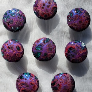 May include: Eight round decorative cabinet knobs with a marbled design. The knobs feature a mix of purple, red, and teal colors, with a glossy finish. The knobs are arranged in a circular pattern on a white surface.