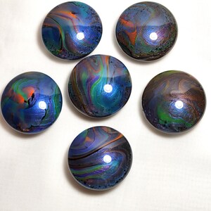 May include: Six circular cabinet knobs featuring a glossy, poured paint finish with swirling patterns of deep blue, vibrant green, bright orange, and purple. Each knob has a smooth, reflective surface with a distinct, unique marbled color blend.