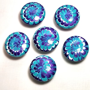 May include: Six circular buttons featuring a vibrant marbled pattern in shades of deep purple, bright turquoise, and white. Each button displays a unique, swirling floral-like design created with glossy paint, arranged in a cluster on a white surface.