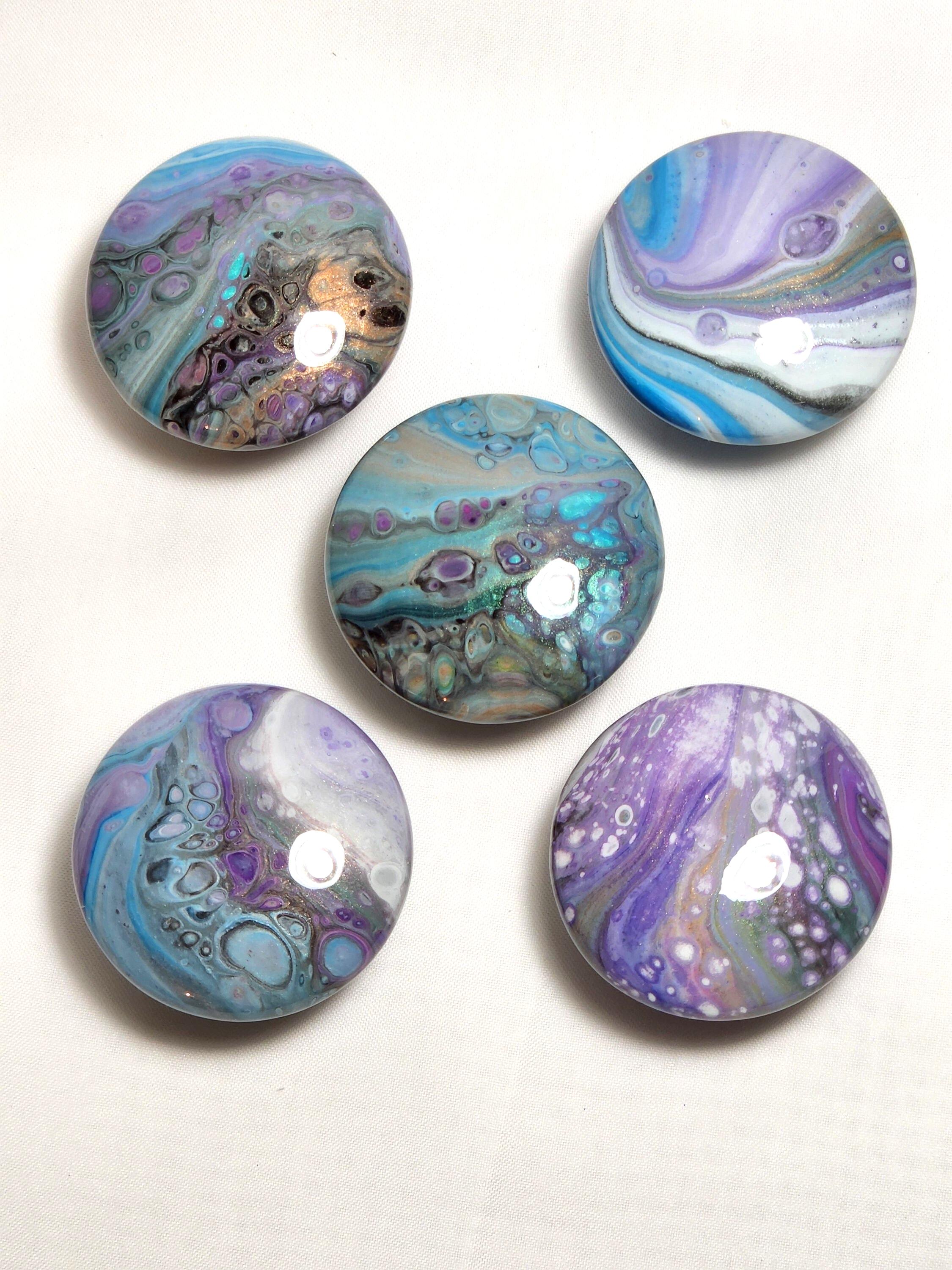 May include: Five circular cabinet knobs featuring a glossy finish with fluid acrylic paint designs. Each knob displays a unique pattern of swirling colors including shades of blue, purple, white, and metallic gold, creating marbled effects with distinct cell-like structures.