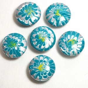 May include: A set of six circular buttons featuring a marbled swirl pattern in shades of teal, silver, and bright yellow. Each button displays a unique fluid design created by the blending of these three colors across the smooth, round surface.