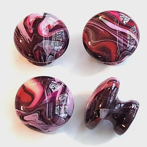 May include: Four round cabinet knobs featuring a swirling marbled pattern in shades of deep burgundy, bright pink, and white, accented with small flecks of metallic silver foil embedded within the smooth, glossy, rounded surface of each knob.