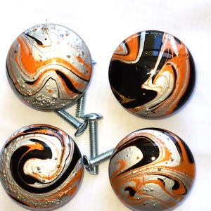 May include: Four round cabinet knobs featuring a glossy, marbled swirl pattern in black, bright orange, and metallic silver. Each knob has a smooth, domed surface and includes a threaded metal mounting screw attached to the back for installation.