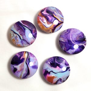 May include: Five circular cabochons featuring a fluid acrylic pour design with swirling patterns of deep purple, light lavender, white, and metallic gold. Each piece has a smooth, glossy, domed surface that reflects light, creating a marbled effect.