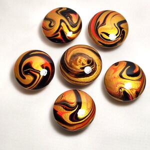 May include: Six circular cabinet knobs featuring a glossy, marbled finish with swirling patterns of metallic gold, deep black, and vibrant orange. Each knob has a smooth, rounded surface that reflects light, creating a polished appearance.