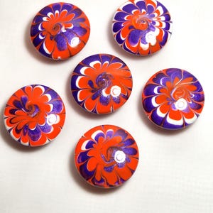 May include: A set of five circular cabinet knobs featuring a vibrant swirl pattern of bright orange, deep purple, and white colors. Each knob has a smooth, glossy finish with a distinct marbled design that radiates from the center outward.