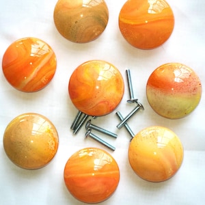 May include: A collection of seven round cabinet knobs featuring a glossy, marbled finish in shades of orange, yellow, and cream. Each knob has a smooth, domed surface and is accompanied by a silver-toned metal mounting screw for installation.