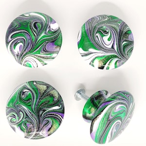 May include: Four round cabinet knobs featuring a swirling marbled pattern in shades of green, purple, black, and white with integrated glitter accents. Each knob has a smooth, glossy finish and includes a silver-toned metal mounting screw.