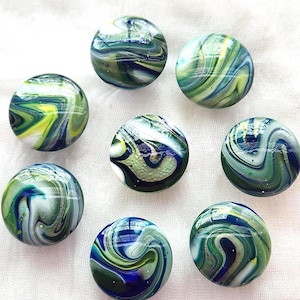 May include: Eight round glass cabochons featuring a swirling marbled pattern in shades of deep blue, forest green, bright yellow, and white. Each piece has a smooth, polished, and reflective surface with unique, organic, and flowing color transitions.