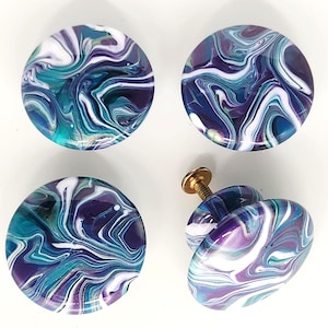 May include: Four round cabinet knobs featuring a swirling marbled pattern in shades of deep purple, teal, white, and dark blue. Three knobs are shown from a top-down perspective, while one knob is positioned on its side to reveal a brass-colored screw.