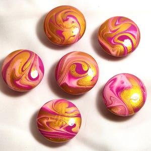 May include: Six circular cabinet knobs featuring a swirling marbled pattern of metallic gold, vibrant magenta, and soft light pink. Each knob has a smooth, glossy finish with unique fluid paint designs that create a distinct look for every piece.
