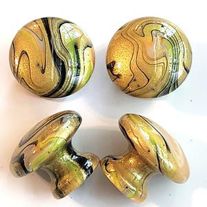 May include: Four round cabinet knobs featuring a swirling marbled pattern in shades of metallic gold, lime green, and black. The knobs have a smooth, glossy finish with a rounded top and a tapered base, displaying a consistent fluid paint design.