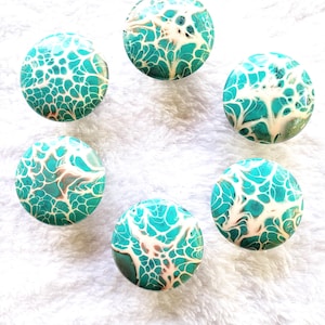 May include: A set of five circular cabinet knobs featuring a fluid acrylic pour design with a marbled pattern of vibrant turquoise, white, and subtle brown veins. Each knob has a smooth, rounded surface with a glossy finish and a consistent pattern.