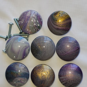 May include: A collection of eight round cabinet knobs with a glossy, marbled finish. The knobs feature swirls of purple, gray, and gold, with some knobs having a speckled appearance. Several silver screws are also visible, suggesting these are ready for installation.